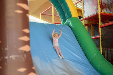 Genç bir çocuk, kapalı bir oyun alanında canlı renklerle çevrili şişirilebilir bir slayttan neşeyle kayıyor. Atmosfer heyecan ve kahkaha ile dolu..