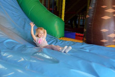 Neşeli genç bir kız, kapalı bir oyun merkezinde canlı, şişme bir kayaktan kayıyor. Heyecan içinde elini kaldırır ve eğlenceli macerasının tadını çıkarır..