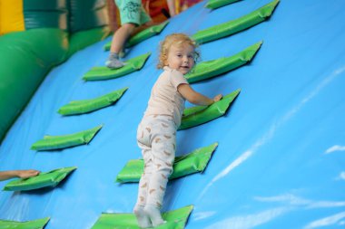 Kıvırcık saçlı bir çocuk enerjik bir şekilde şişme duvara tırmanıyor. Çocuklar oyun sahasında eğlenirken neşeli ortam heyecanla uğulduyor..