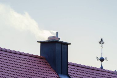 Aydınlık bir günde, duman bir evin bacasından hafifçe süzülür. Çatıda belirgin kırmızı fayanslar, ve şık bir rüzgar gülünün tepesi sahneye cazibe katıyor..