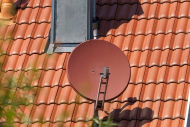 Bir uydu anteni terracotta çatısına kurulur, açık bir günde güneş ışığını yakalar. Çanak, manzarayı yeşillendiren bir yerleşim alanına yerleştirilmiştir..