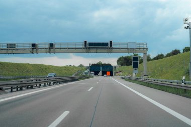 Araçlar otoyoldaki bir gişeye doğru yol alıyorlar. Bulutlu gökyüzü, akşam erken saatlerde, arazi yeşil ve yol kenarında yemyeşil kaldığı için bastırılmış bir atmosfer yaratır..