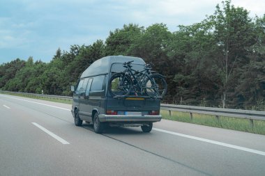 Koyu renkli bir minibüs otoyol boyunca gidiyor, arkada bir askıda iki bisiklet taşıyor. Sahnede bulutlu bir gökyüzünün altında bir yeşillik arkaplanı yer alıyor..