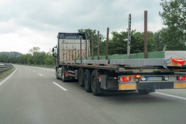 Büyük bir kamyon, bulutlu bir gökyüzünün altındaki yemyeşil otobanda inşaat malzemeleri taşıyor. Olay yerinde altyapı ve lojistik faaliyet gösteriyor.