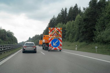 Otoyolda parlak uyarı işaretleri olan inşaat araçları dizildi. Yeşil ağaçların ve bulutlu gökyüzünün ortasından bir araba yol çalışması alanına yaklaşıyor..
