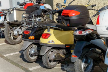 Değişik renklerde çeşitli scooterlar canlı bir şehir ortamında birbirine yakın park edilir ve günlük ulaşım seçenekleri gösterilir. Güneşli hava canlı atmosferi güçlendirir..