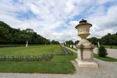 Ziyaretçiler zarif vazoların, yemyeşil çiçeklerin ve bulutlu bir gökyüzünün altındaki klasik heykellerin yer aldığı titizlikle manikürlü bir bahçede geziniyorlar..