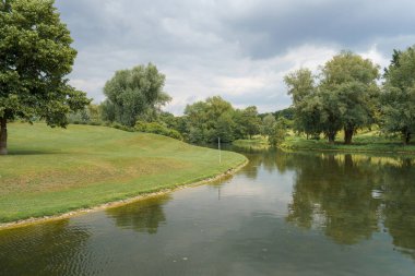 Sakin bir nehir kıyısında yeşil çimlerle kaplı yumuşak yamaçlar ve suyu kaplayan ağaçlar bulunur. Gökyüzü bulutlu, öğleden sonra saatlerinde huzurlu bir atmosfer yaratıyor..