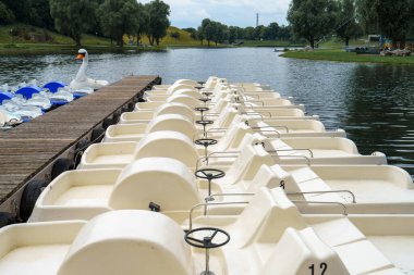 Sakin bir göl parkındaki ahşap rıhtımda parlak kürek tekneleri bekliyor. Sakin sular yemyeşil ve berrak gökyüzünü yansıtarak ziyaretçiler için davetkar bir atmosfer yaratıyor..
