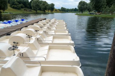 Bereketli yeşilliklerle çevrili sakin bir gölün kıyısında bir sıra kürek kayığı duruyor. Bulutlu gökyüzü öğleden sonra aktiviteleri için sakin bir atmosfer yaratır..