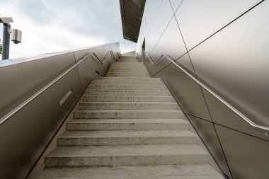 Beton merdivenler kentsel bir ortamda şık bir bina girişine doğru yükseliyor. Mimari düz metalik yüzeylere ve bulutlu bir gökyüzüne karşı modern bir tasarıma sahiptir..