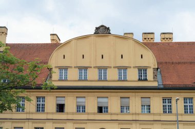 Tarihi bina dekoratif unsurlarla çarpıcı bir sarı cephe içeriyor. Pencereler üst katta eğri bir çatı altında, bulutlu bir gökyüzünün arka planına göre dizilmiş..
