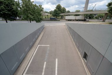 Yoğun bir yola bağlanan ve geçen kamyonları olan bir şehir altgeçidi manzarası. Ağaçlar bulutlu bir gökyüzünün altındaki bölgeyi çiziyor. Bu da gündüz faaliyetlerini gösteriyor..