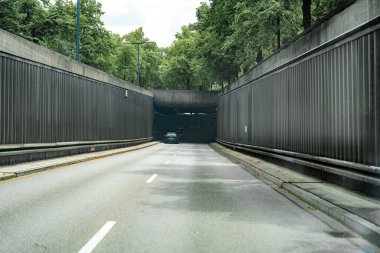 Bir araba, uzun ağaçlarla çevrili karanlık bir tünel girişine doğru gidiyor ve gün ışığında doğa ile şehir altyapısı arasında bir zıtlık oluşturuyor..