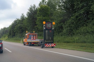Otoyola parlayan ışıklarla bir hız limiti tabelası konurken, öğleden sonra yakınlarda bir servis aracı park halinde bulunuyor..