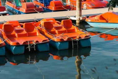 Parlak turuncu pedallı botlar bir marinaya yanaşır ve durgun suda yansımalar oluştururlar. Açık, güneşli bir gün, eğlence ve eğlence için ideal..