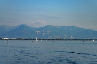 Yelkenliler, uzak dağlarla çevrili Cenevre Gölü 'nün sakin sularında yol alırlar. Açık gökyüzü ve yumuşak dalgalar güneşli bir öğleden sonra huzurlu bir atmosfer yaratır..