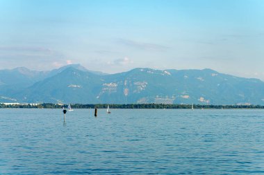 Yelkenliler, güneşli bir gökyüzünün altındaki görkemli dağların çerçevelediği berrak mavi bir gölde süzülüyor. Huzurlu su manzarayı yansıtır. Rahatlamayı davet eder..