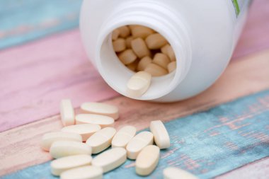 Oval beyaz tabletler pastel renkli ahşap bir yüzeye saçılmış, bazıları da ters dönmüş plastik bir şişede kalmış. Ayar parlak ve davetkar, canlı bir karşıtlık oluşturuyor..