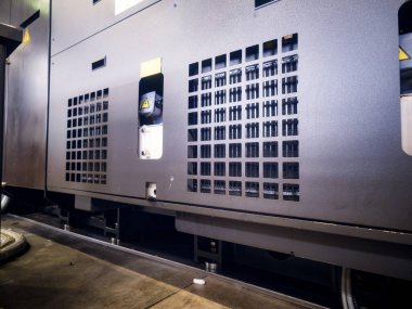 Karanlık bir sanayi bölgesi, birden fazla havalandırma ızgarası olan büyük bir makinenin yan görüntüsünü gösteriyor. Makineler sürekli çalışmayı sağlarken ışıklar çevreyi aydınlatır..