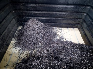 Koyu metal talaşları büyük siyah bir konteynırda toplanıyor. Parlak doğal ışık ile dolu işlek bir atölyedeki işlemin kalıntılarını sergiliyorlar..