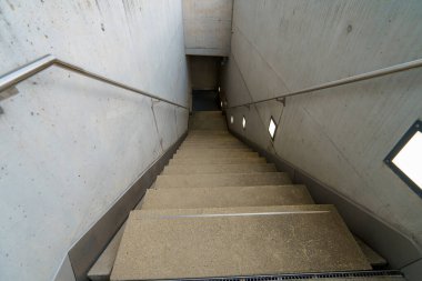 Duvarlar boyunca uzanan küçük ışıklarla aydınlatılmış dar bir koridora beton bir merdiven iner. Tasarım modern, temiz çizgiler ve minimalist estetik..