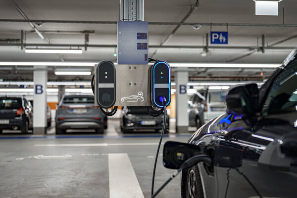 Электростанция зарядки транспортных средств расположена в просторной подземной автостоянке. Несколько автомобилей, некоторые с зарядными кабелями, припаркованы неподалеку, демонстрируя современную инфраструктуру.