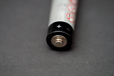 Bir batarya karanlık bir yüzeye dayanır, metal pozitif terminalini vurgular ve kaplamasına çeşitli güvenlik uyarıları basılır, işlevsel tasarımı resmedilir.