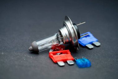 Bir halojen ampulü karanlık bir yüzeyde renkli fünyelerin yanında duruyor ve önemli otomotiv bileşenlerini vurguluyor. Bu parçalar araç bakımı ve işlevselliği için gereklidir, tamir çalışmaları için uygundur..