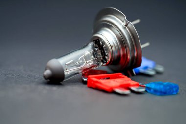 Bir araba farının ampulü karanlık bir yüzeyde uzanıyor, şeklini ve iç bileşenlerini gösteriyor. Yakınlarda, kırmızı ve mavi renkli elektrik konnektörleri, otomotiv tamir ögelerini gösteren bağlam sağlar.