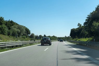 Vehicles travel along a smooth highway lined with lush greenery. The bright blue sky indicates a warm summer day perfect for a road trip.