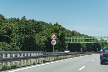 A speed limit sign showing 120 km/h is visible on a highway. The scene features clear blue skies with a few clouds and greenery lining the roadside. Vehicles travel along the road.