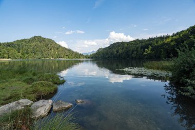 A tranquil lake surrounded by lush green hills reflects the blue sky and white clouds. The scene captures a peaceful moment in nature on a sunny day.