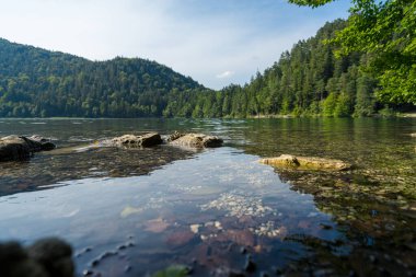 A serene lake scene features smooth water reflecting surrounding green mountains. Rocks are visible along the water's edge, adding to the tranquility of the natural setting.