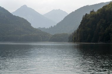 Still waters reflect the gray sky at a tranquil mountain lake. Lush green hills rise on either side, with distant peaks shrouded in mist. A few boats navigate the surface.