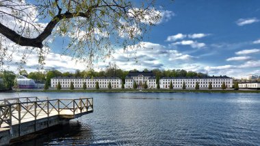 Stockholm 'deki Karlbergs Sarayı' nı ve açık mavi bir bahar gökyüzünün altındaki rıhtımı gösteren geniş bir liman manzarası..
