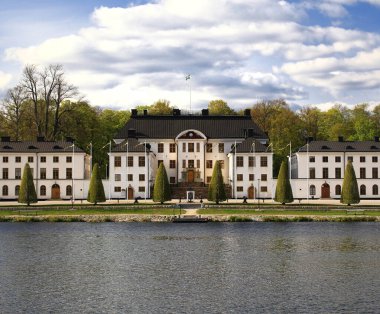 Stockholm 'deki Beyaz Tarihi Karlbergs Sarayı simetrik bir manzarada bayrak ve ağaçlarla dolu sakin sulara sahip..