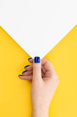 A womans hand with a blue manicure holds an empty sheet of paper on a yellow background, top view, copy space.