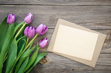 Bouquet of purple tulips and blank greeting card on wooden background, top view, copy space.