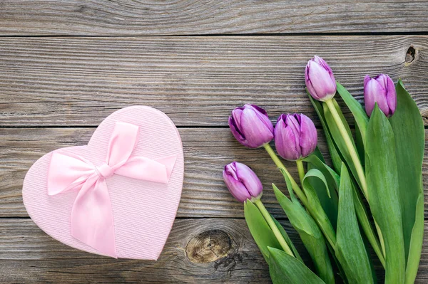 A bouquet of purple tulips and a heart-shaped gift box, on a wooden background, top view.
