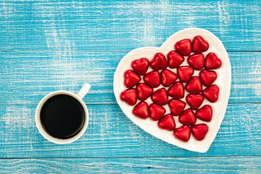 Heart shaped plate with heart shaped red foil chocolates and cup of coffee on blue wooden background, top view, Valentines day concept.