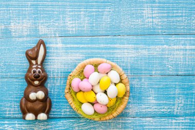 Easter background with chocolate bunny and colorful chocolate eggs in a nest on a blue wooden background, flat lay, Easter holiday concept, childrens sweets, copy space.