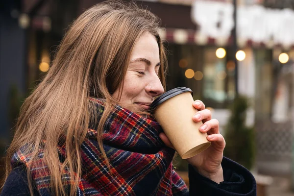 Stylish young woman with a nose piercing drinks coffee from a paper cup on a walk in the city, blurred background with bokeh, urban portrait in the cold season.