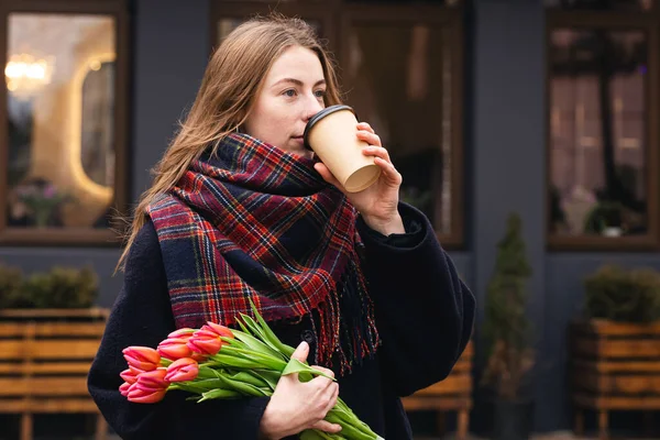 Stylish young woman with tulips bouquet drink coffee outdoors, spring street lifestyle portrait.