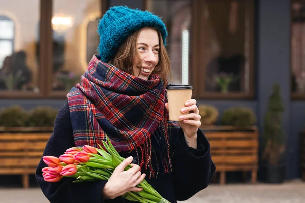 Stylish young woman with tulips bouquet and coffee outdoors, spring street lifestyle portrait.