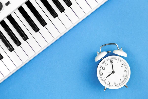 White alarm clock and piano on a blue background, top view, the concept of music education and creativity, white synthesizer, flat lay.