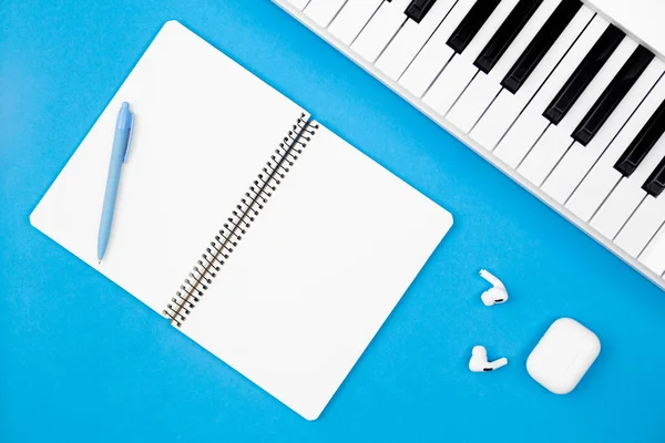 Blank notebook and piano on blue background, top view, concept of musical creativity and education, white synthesizer, flat lay.
