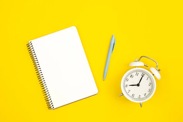 stock image White alarm clock and blank notepad on yellow background, flat lay, copy space.