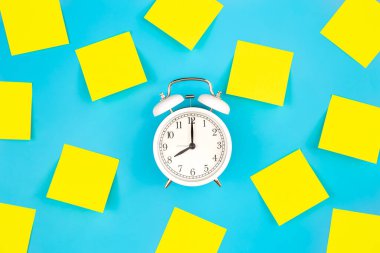 Beyaz alarmlı saat ve sarı kağıt yakında geliyor. Mavi arka planda boş sarı yapışkan notlar. Zaman yönetimi, öncelikler, verimlilik, kontrol ve hedefler kavramı.