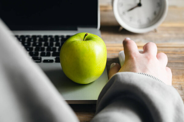 Женщина работает за ноутбуком, яблочный крупный план, будильник на заднем плане, концепция здорового завтрака и закуски на работе.